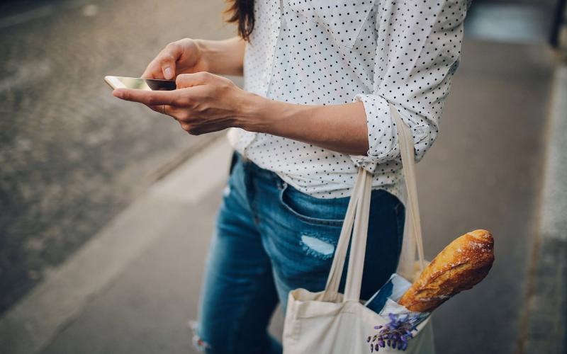 woman walks down the street and texts with a bag of groceries on her arm